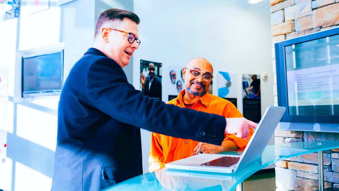 Two smiling male colleagues pointing at laptop on standing desk