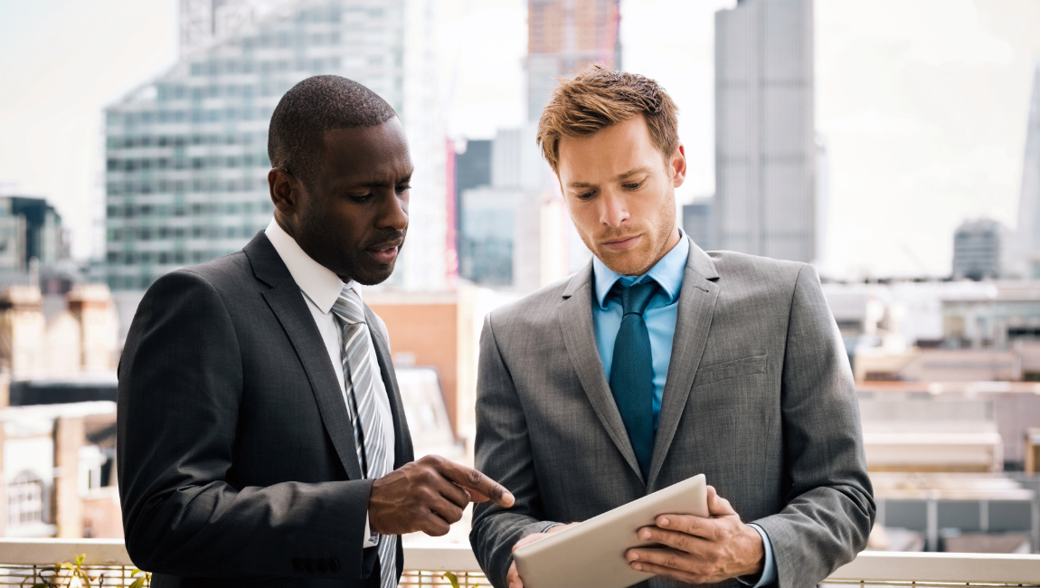 Two men working together pointing at tablet while standing on office terrace downtown